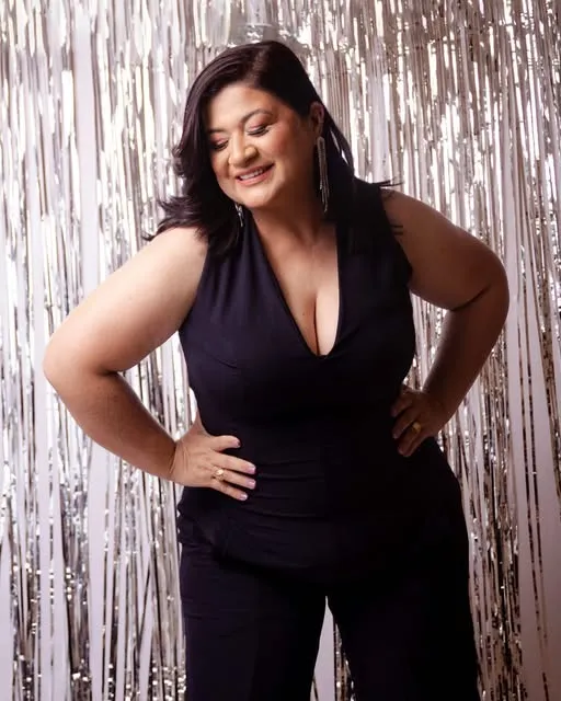 Sandra sorrindo, com as mãos na cintura, em um look preto diante de um fundo prateado
