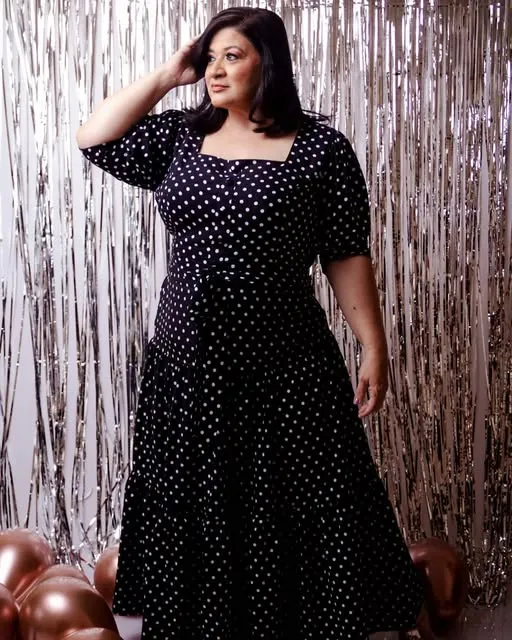 Sandra usando um vestido preto de bolinhas brancas, diante de um fundo prateado